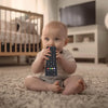 Baby sitting on carpet chewing a silicone teether toy shaped like a TV remote control.